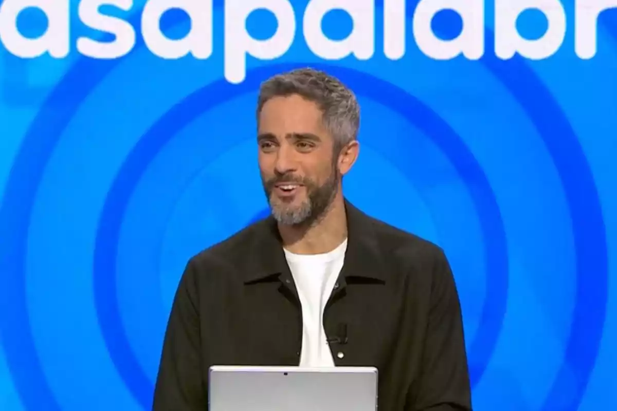 Roberto Leal con barba y cabello corto sonríe mientras está de pie frente a un fondo azul con letras blancas.