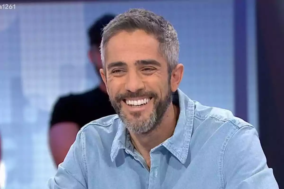 Un home somrient amb barba i camisa texana en un estudi de televisió.