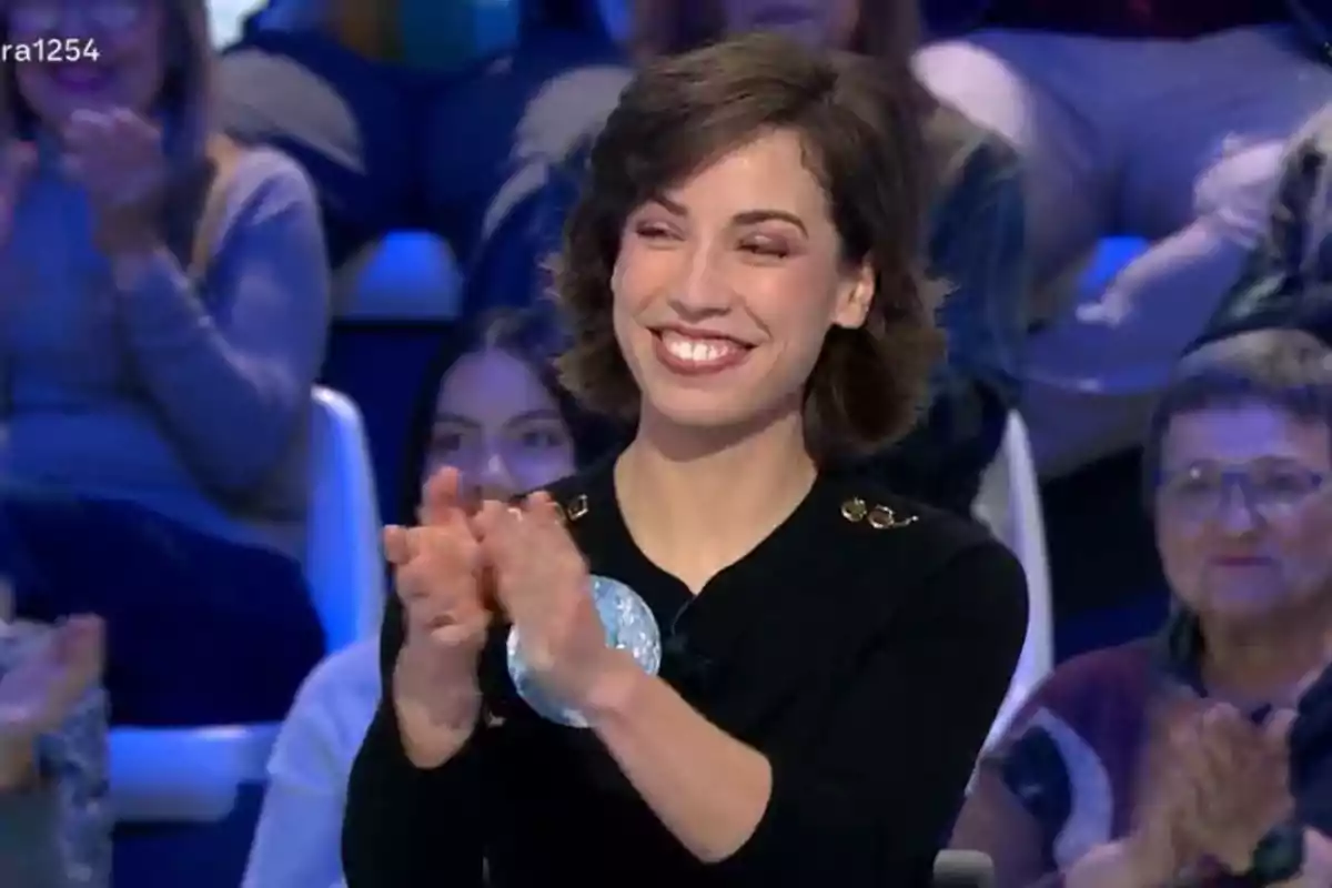 Una dona somrient aplaudeix en un programa de televisió amb públic al fons. Una dona somrient aplaudeix en un programa de televisió amb públic al fons.