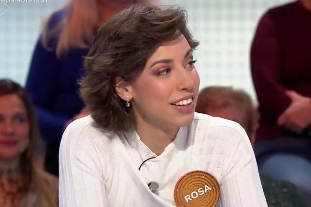 A woman with short brown hair smiles while participating in a television show, wearing a white sweater and a brooch with the name A woman with short brown hair smiles while participating in a television show, wearing a white sweater and a brooch with the name