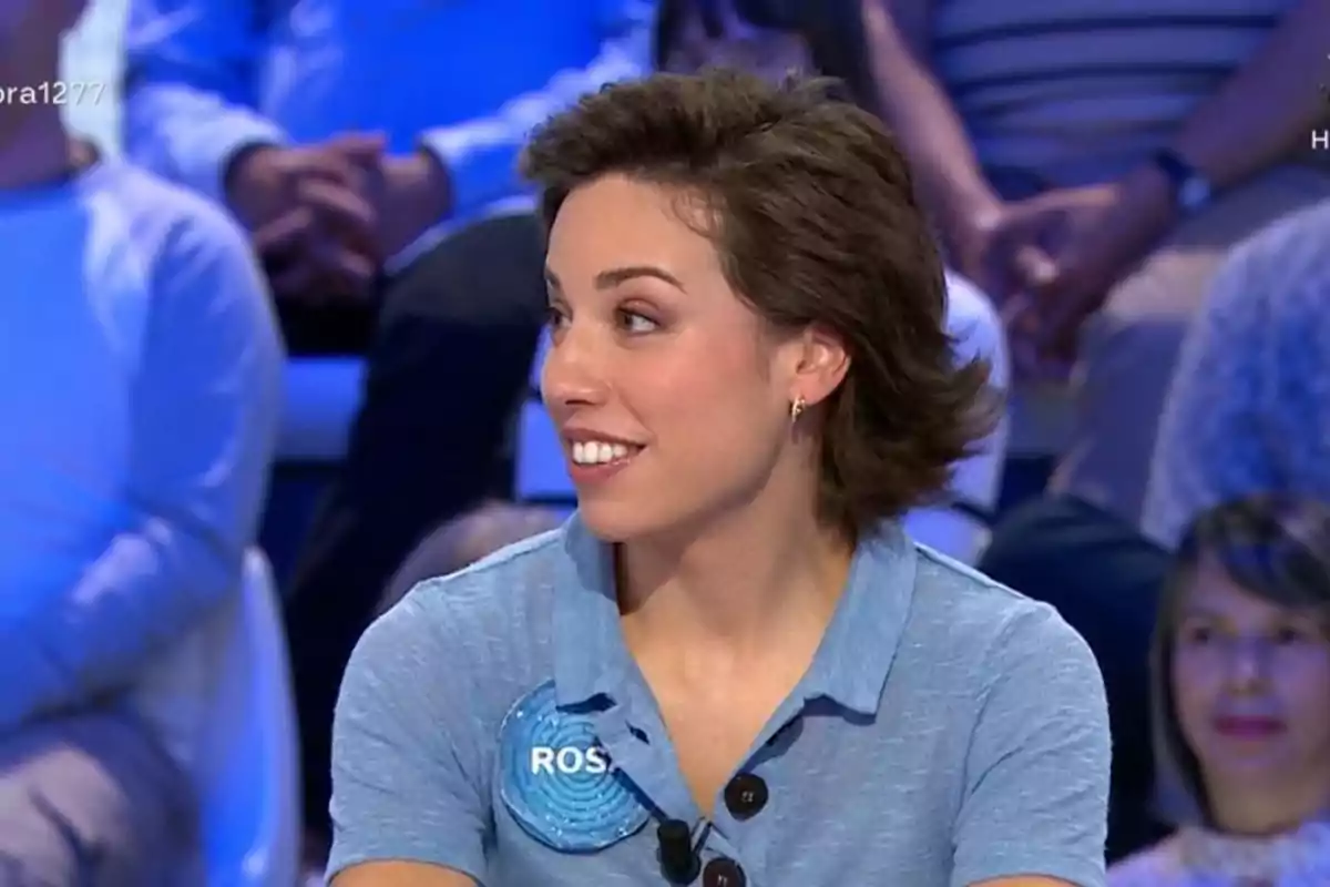 Dona jove amb cabell curt i castany, vestida amb una camisa blau clar i una placa amb el nom "ROS", asseguda en un plató de televisió amb públic al fons