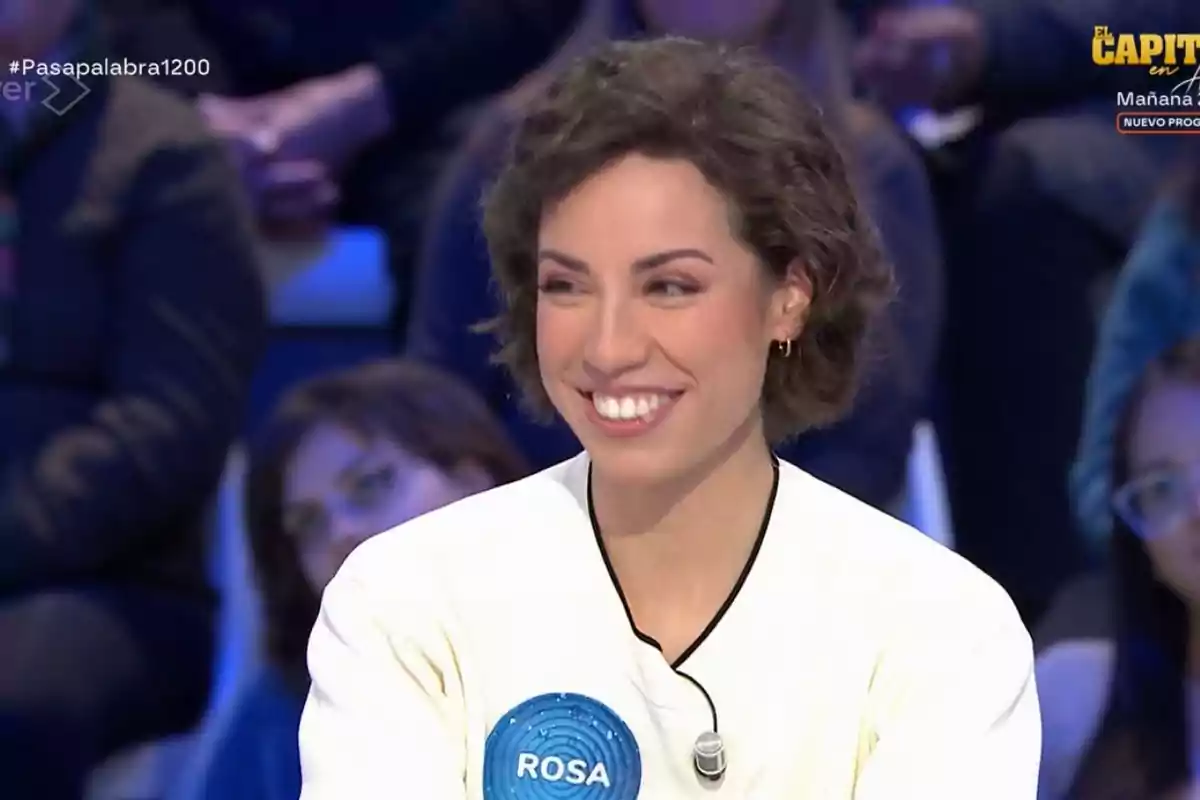 Una persona somrient amb cabell curt i arrissat, porta una samarreta blanca amb un micròfon de solapa i un cercle blau amb el nom Una persona somrient amb cabell curt i arrissat, porta una samarreta blanca amb un micròfon de solapa i un cercle blau amb el nom