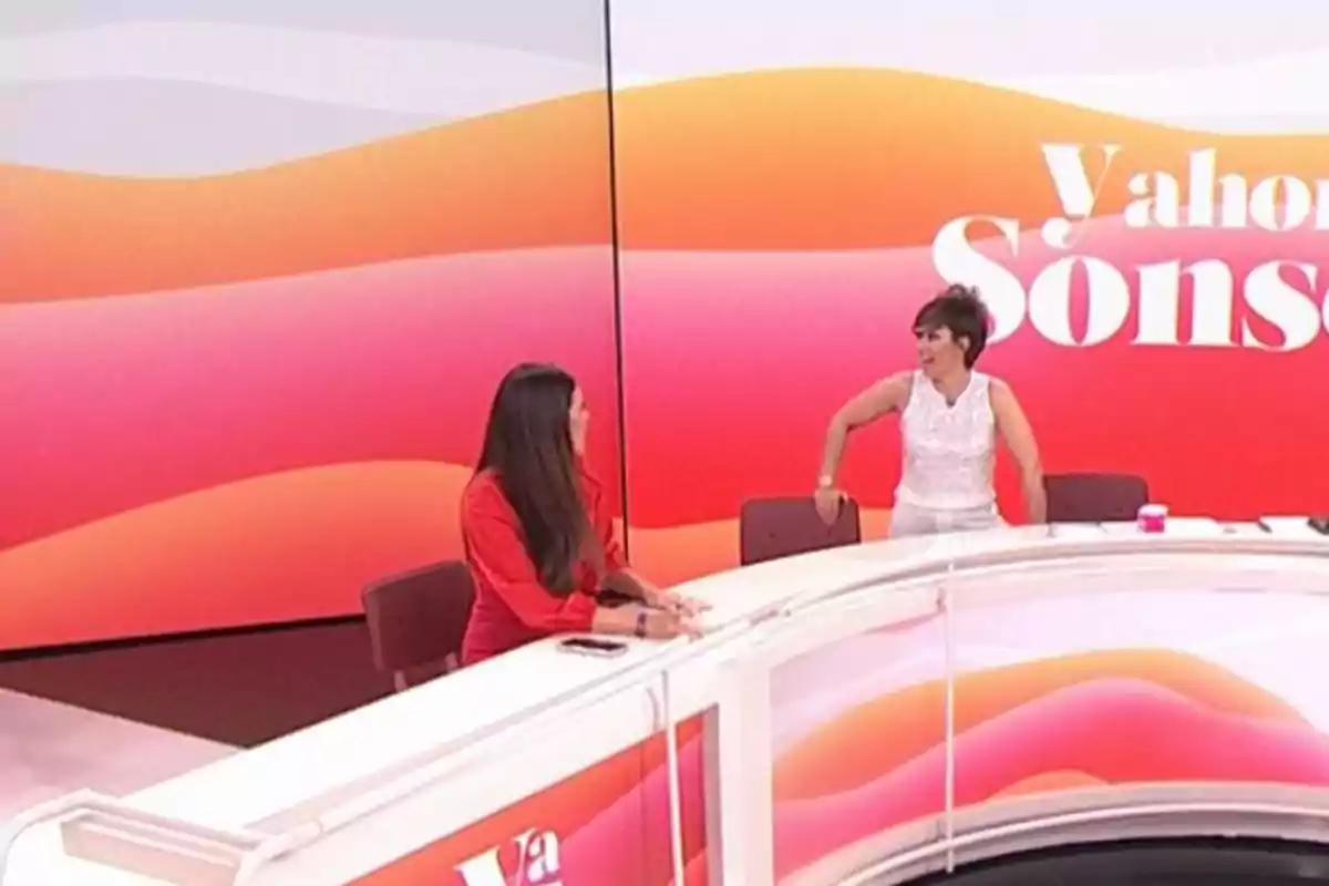 Dues persones conversen en un plató de televisió amb un fons acolorit i un escriptori corbat. Dues persones conversen en un plató de televisió amb un fons acolorit i un escriptori corbat.