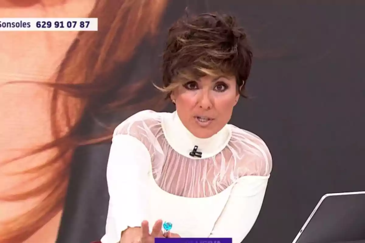 Sonsoles Ónega con cabello corto y oscuro está hablando en un programa de televisión, lleva una blusa blanca con detalles transparentes y hay un número de teléfono en la esquina superior izquierda.