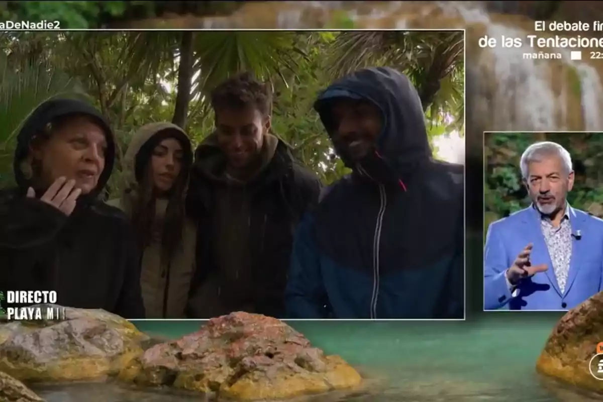 A group of people in warm clothing is in a jungle setting while an older man appears in a box speaking in a studio. A group of people in warm clothing is in a jungle setting while an older man appears in a box speaking in a studio.