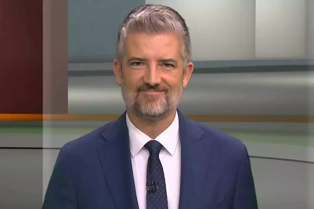 Un home amb barba i cabell canós porta un vestit blau i corbata, somrient davant d'un fons d'estudi. Un home amb barba i cabell canós porta un vestit blau i corbata, somrient davant d'un fons d'estudi.