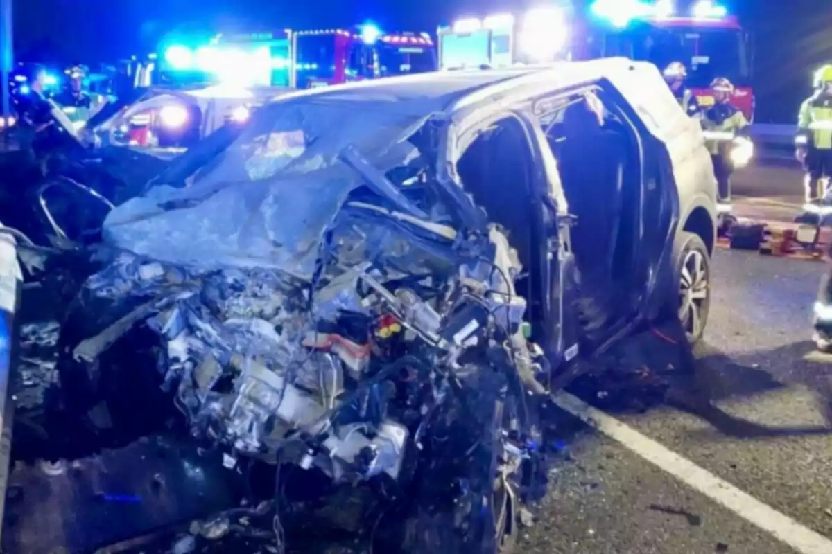 Cotxe greument danyat després d’un accident a la carretera de nit, amb llums d’emergència i personal de rescat al fons