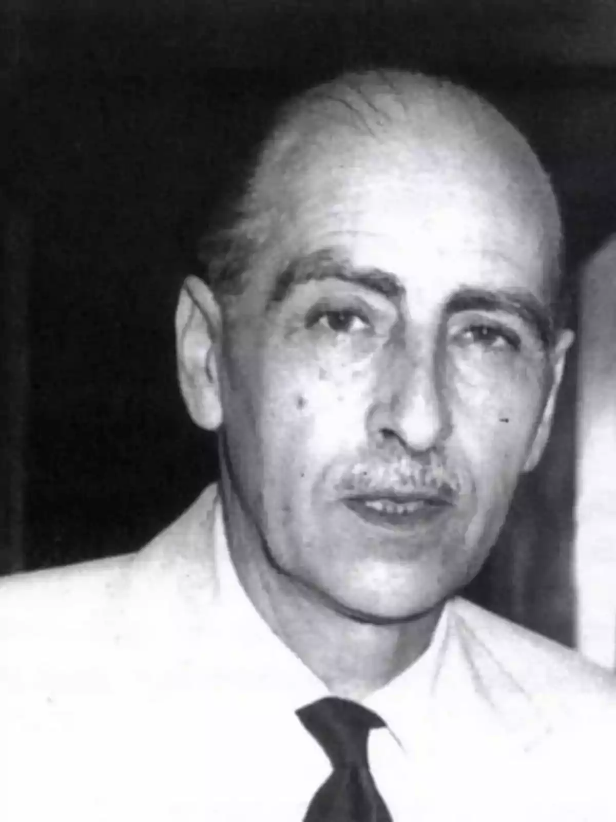 Home de mitjana edat amb bigoti i cabell curt, vestit amb camisa clara i corbata fosca, mirant cap a la càmera en una fotografia en blanc i negre Home de mitjana edat amb bigoti i cabell curt, vestit amb camisa clara i corbata fosca, mirant cap a la càmera en una fotografia en blanc i negre