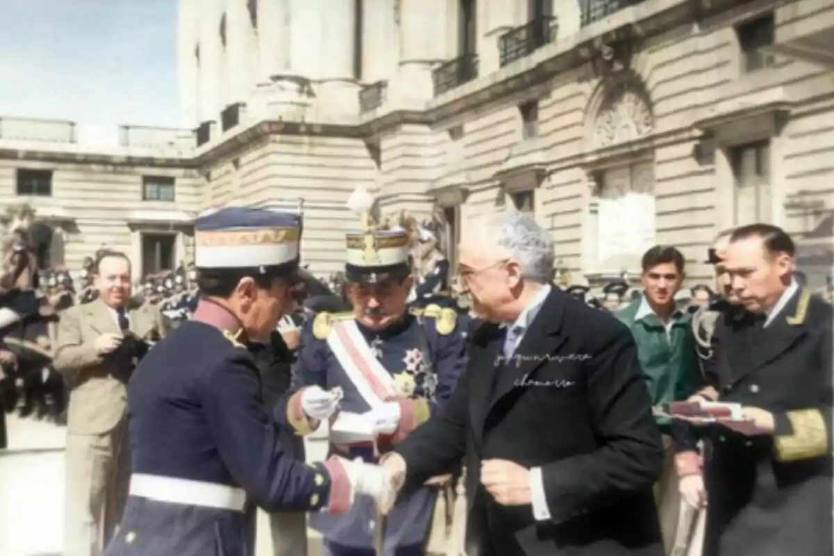 Un grup de persones, algunes amb uniformes militars, es troben en un esdeveniment formal a l'aire lliure davant d'un edifici històric. Un grup de persones, algunes amb uniformes militars, es troben en un esdeveniment formal a l'aire lliure davant d'un edifici històric.