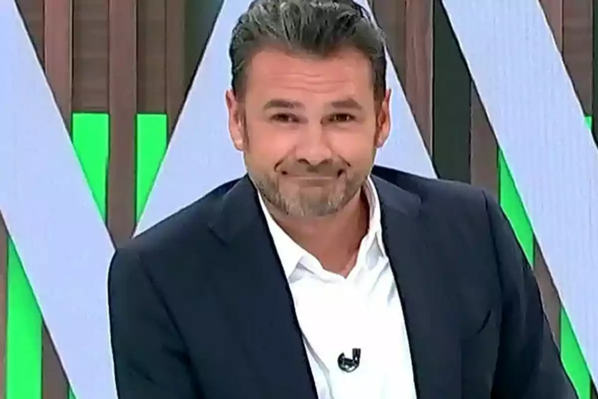 Iñaki López con barba y cabello oscuro, vestido con un traje oscuro y camisa blanca, sonríe ligeramente mientras está de pie frente a un fondo con patrones geométricos y colores verde y marrón.