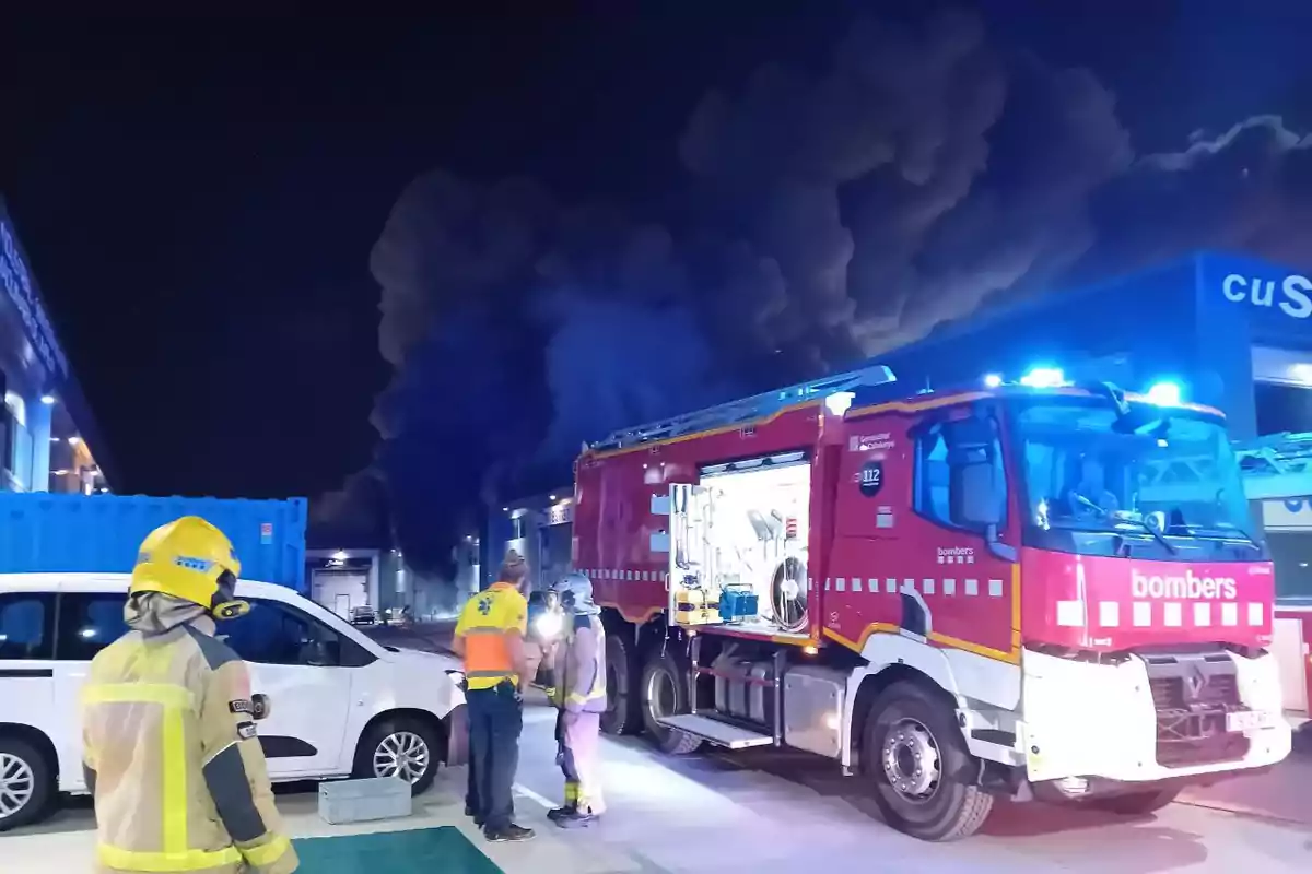 Bombers i personal d'emergència treballant a l'escena d'un incendi nocturn amb un camió de bombers i fum fosc al fons. Bombers i personal d'emergència treballant a l'escena d'un incendi nocturn amb un camió de bombers i fum fosc al fons.