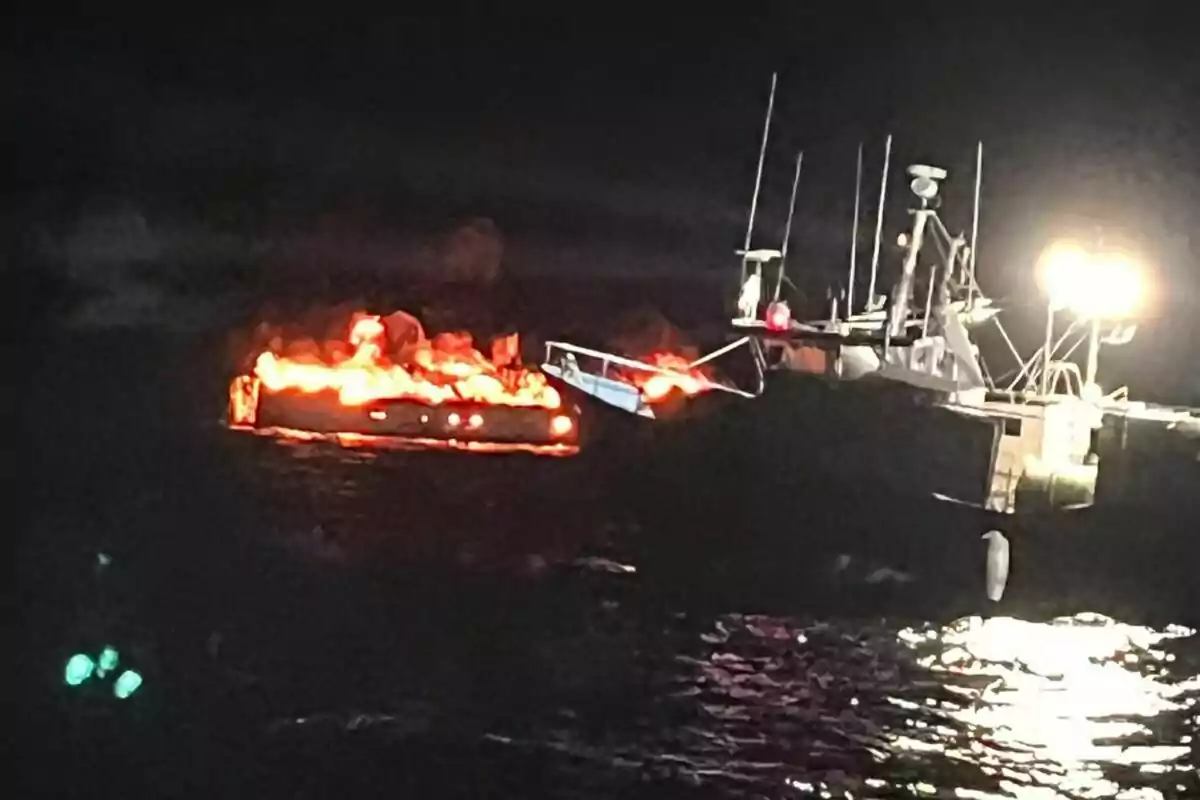 Una embarcació en flames al mar durant la nit amb una altra embarcació il·luminada a prop.