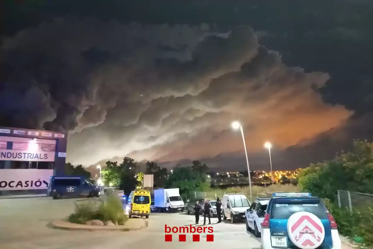 Una escena nocturna mostra un gran núvol de fum fosc sobre una àrea industrial il·luminada per llums d'emergència i vehicles de serveis d'emergència estacionats al lloc.