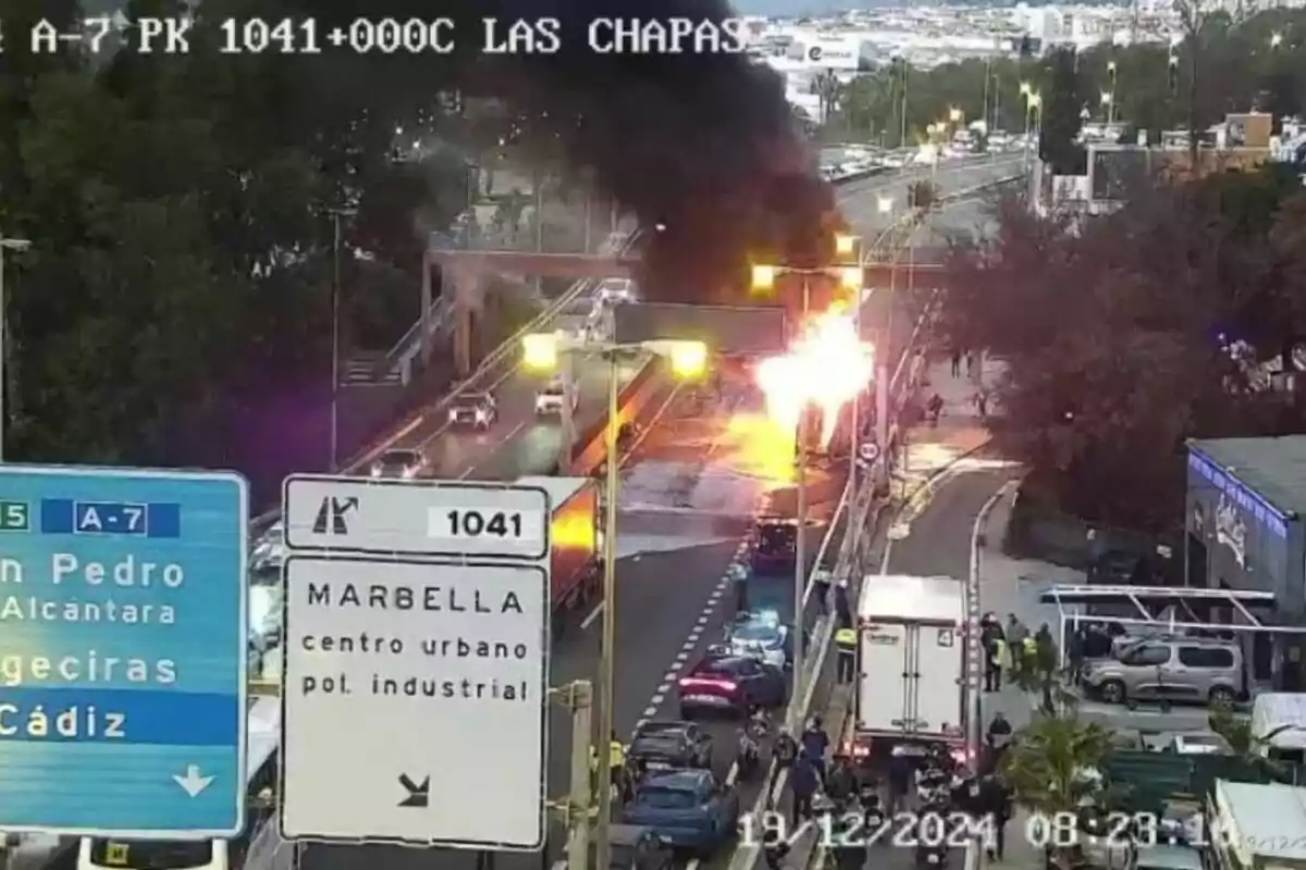 Una carretera con tráfico y un incendio visible en el lado derecho, con señales que indican direcciones hacia Marbella y Cádiz.