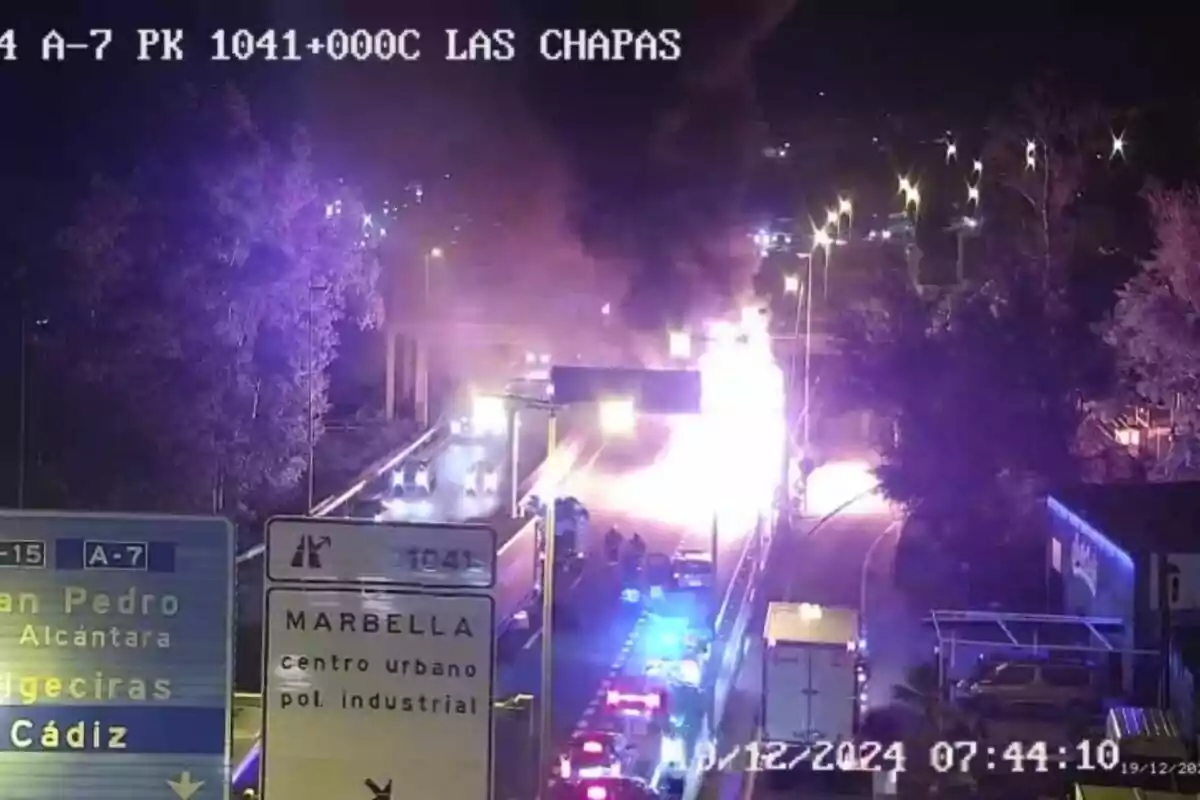 Una carretera de noche con un incendio visible y vehículos de emergencia en la escena, junto a señales de tráfico que indican direcciones hacia San Pedro, Algeciras y Cádiz.