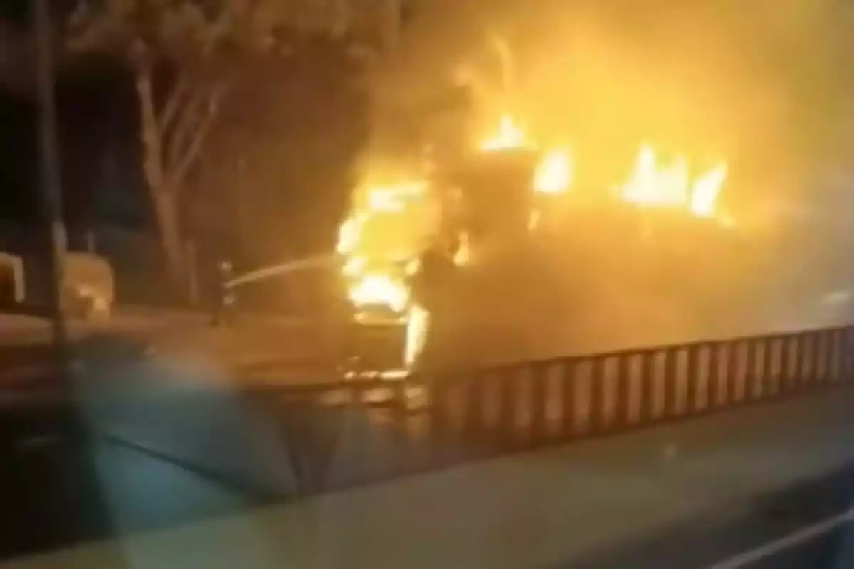 Un vehículo en llamas en una calle durante la noche con un bombero intentando apagar el fuego.