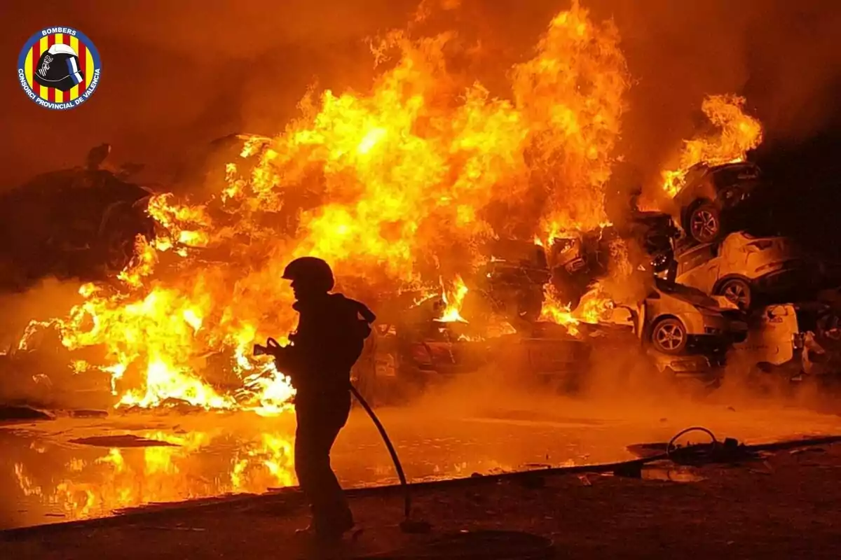 Bombero combatiendo un incendio en un depósito de vehículos con llamas intensas en el fondo.