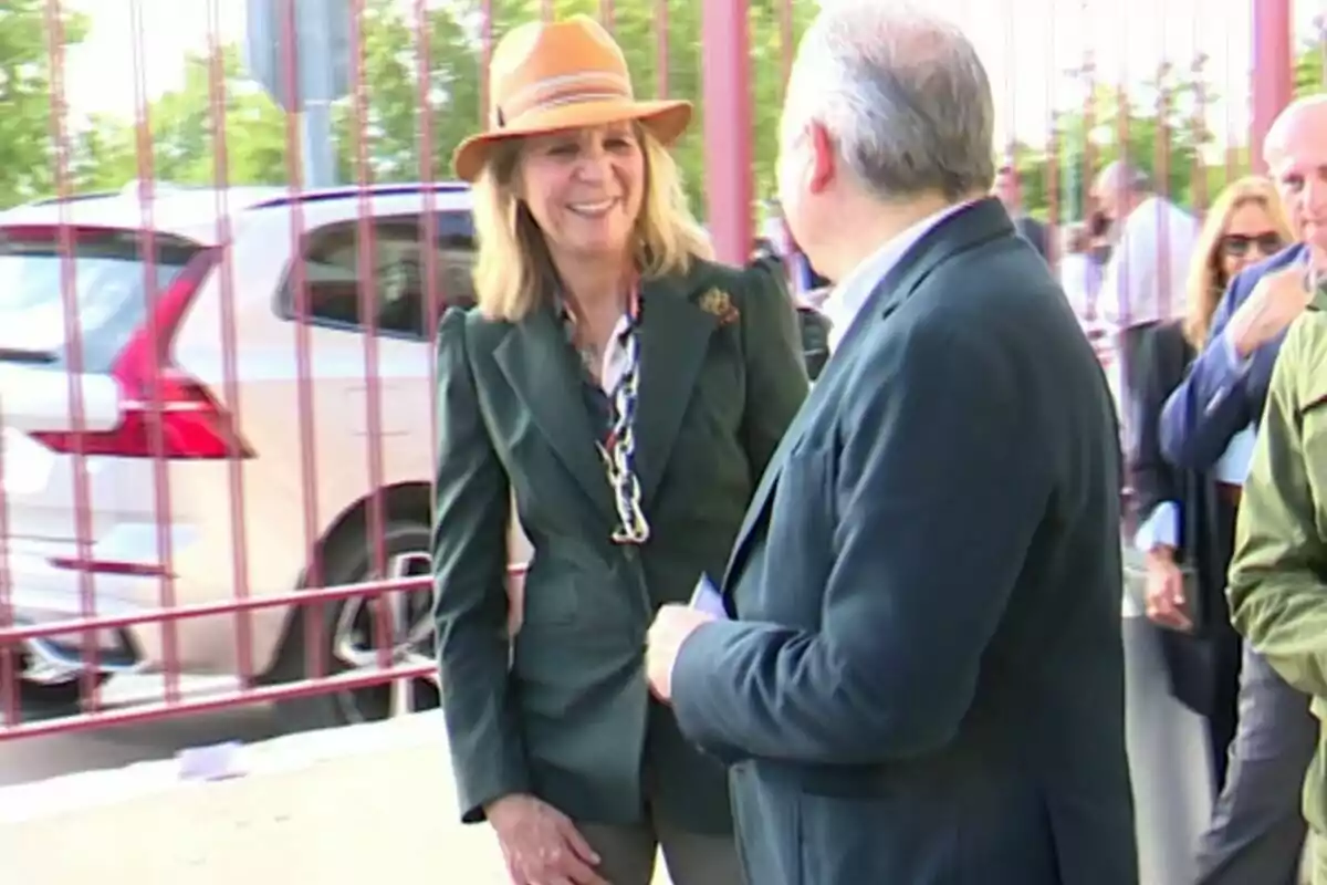 La infanta Elena amb barret somriu mentre conversa amb un home en un entorn exterior amb cotxes i persones de fons. La infanta Elena amb barret somriu mentre conversa amb un home en un entorn exterior amb cotxes i persones de fons.