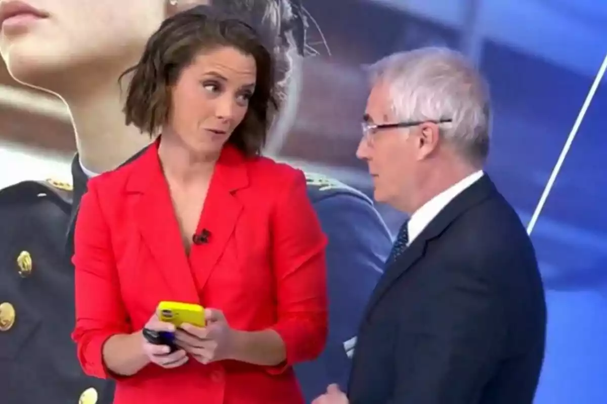 María Casado y David Cantero conversan en Informativos Telecinco; una mujer con chaqueta roja sostiene un teléfono amarillo mientras mira al hombre que lleva traje y corbata.