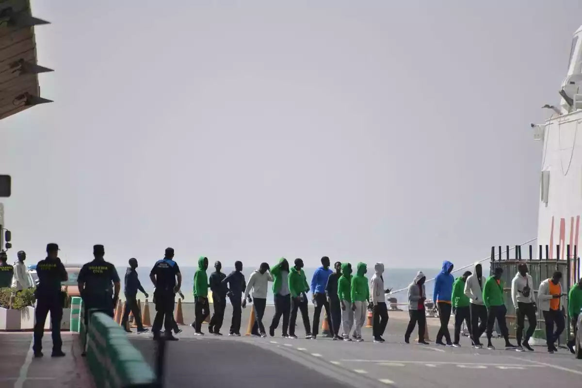 Pla general amb una cua d'immigrants nouvinguts a El Hierro pujant a un vaixell mentre uns Guàrdia Civils els vigilen