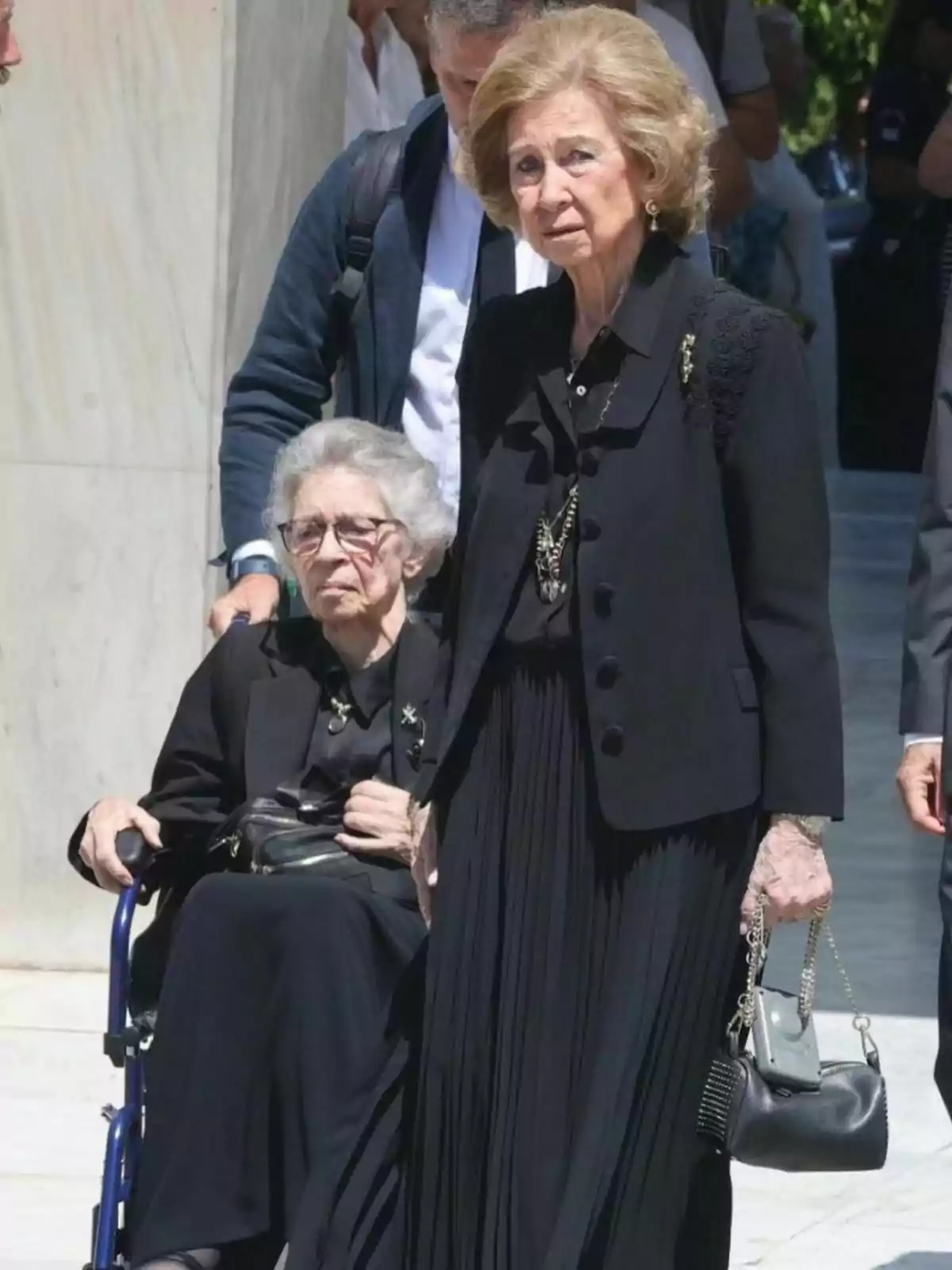 Irene of Greece and Queen Sofia dressed in black, Irene in a wheelchair and Sofia standing holding a bag, with people in the background. Tweet from August 1, 2024