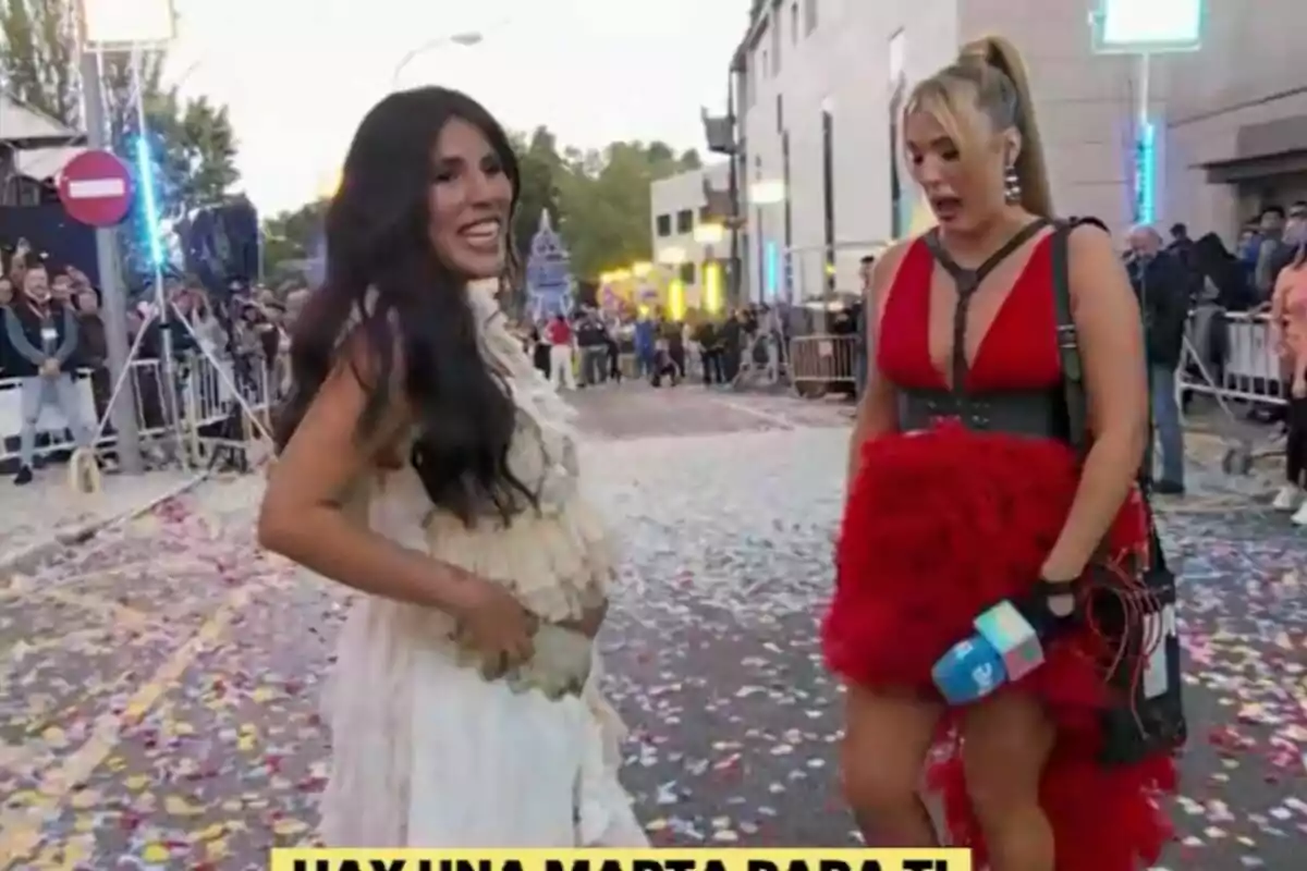 Dues dones són en un carrer ple de confeti, Isa Pantoja porta un vestit blanc i Marta Riesco un vestit vermell, mentre un grup de persones observa des del fons a La Família de la Tele. Dues dones són en un carrer ple de confeti, Isa Pantoja porta un vestit blanc i Marta Riesco un vestit vermell, mentre un grup de persones observa des del fons a La Família de la Tele.