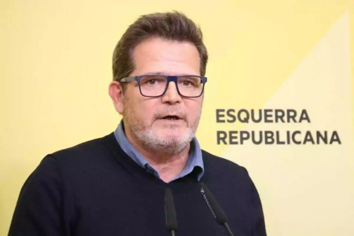Home amb ulleres i barba parlant davant d’un micròfon amb un fons groc on es llegeix Esquerra Republicana Home amb ulleres i barba parlant davant d’un micròfon amb un fons groc on es llegeix Esquerra Republicana