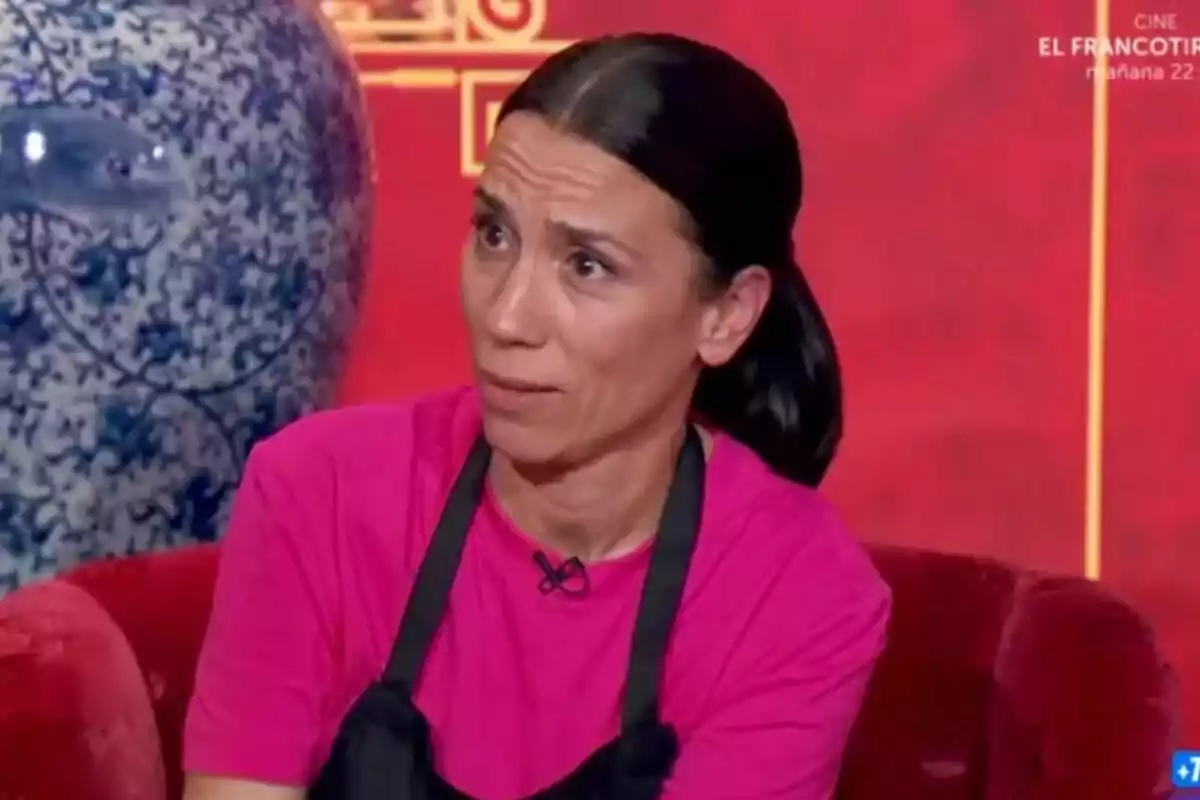 Itziar Miranda amb expressió de sorpresa o confusió, vestida amb samarreta rosa i davantal negre, asseguda en un sofà vermell amb un gerro blau decoratiu al fons a MasterChef Celebrity.