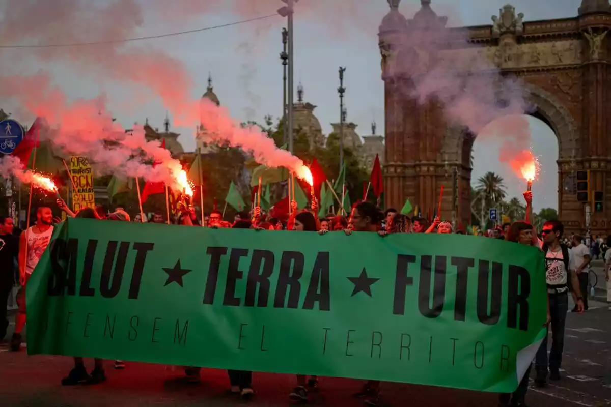 Manifestació amb persones sostenint bengales i una pancarta verda que diu "SALUT TERRA FUTUR" davant de l'Arc de Triomf.