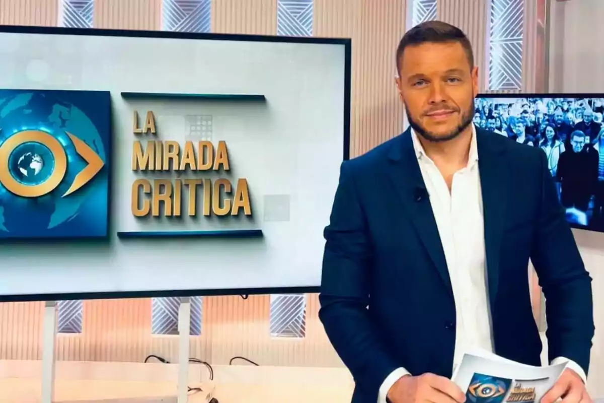 Jano Mecha dret al costat d'una pantalla que mostra el logotip del programa "La Mirada Crítica".