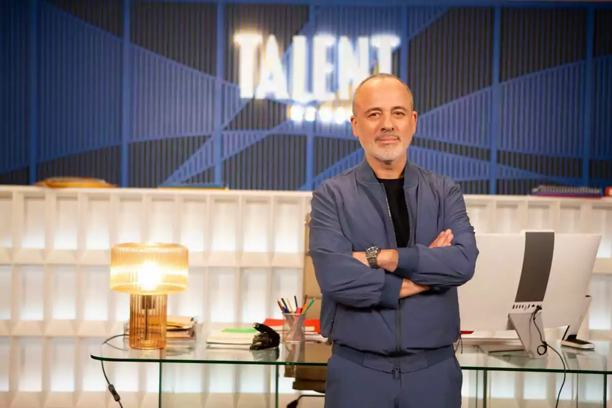 Javier Gutiérrez de pie con los brazos cruzados frente a un escritorio con una lámpara y una computadora en un entorno moderno con un letrero que dice Javier Gutiérrez de pie con los brazos cruzados frente a un escritorio con una lámpara y una computadora en un entorno moderno con un letrero que dice