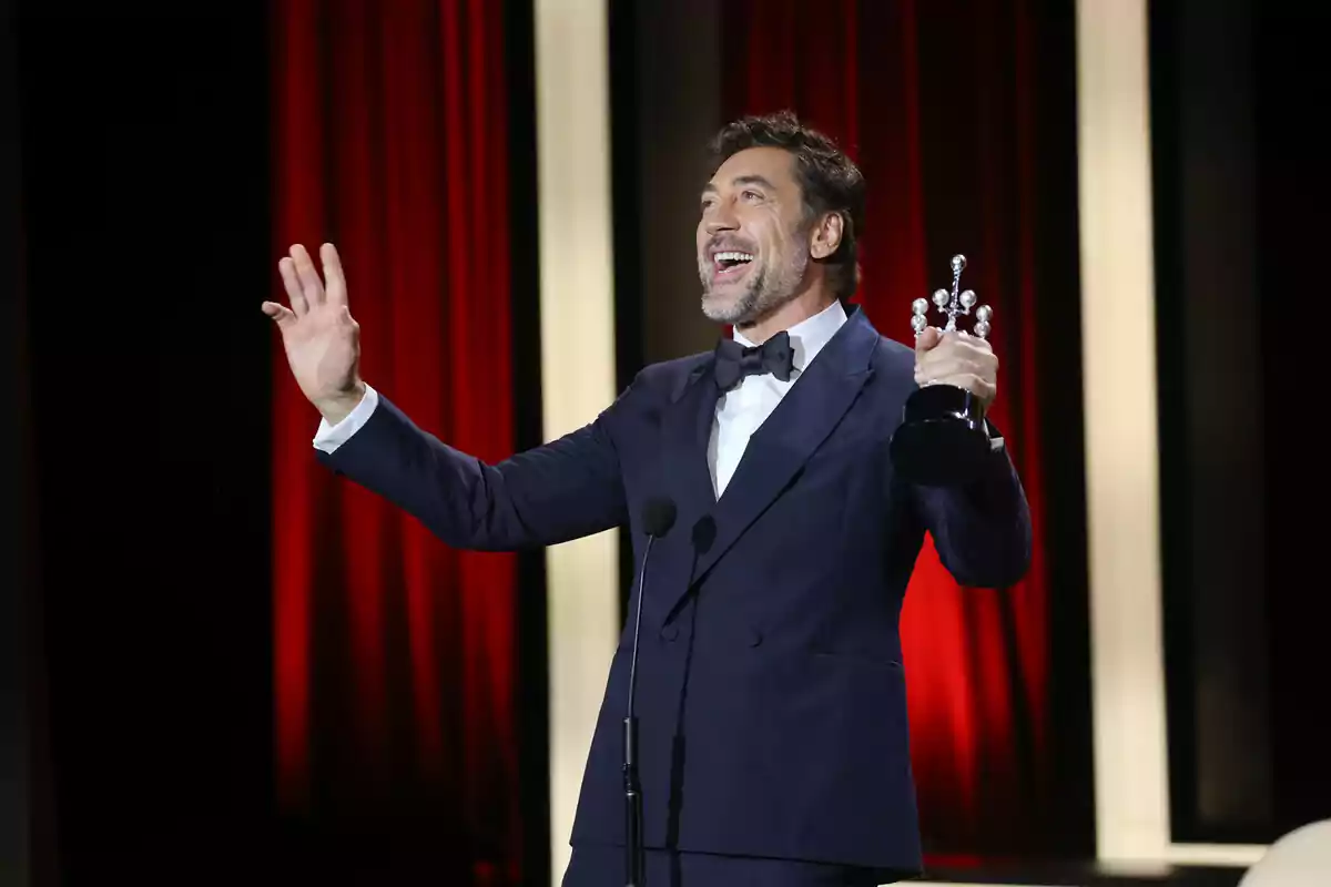 Javier Bardem en un escenario con un traje oscuro sosteniendo un trofeo y sonriendo mientras habla al micrófono.