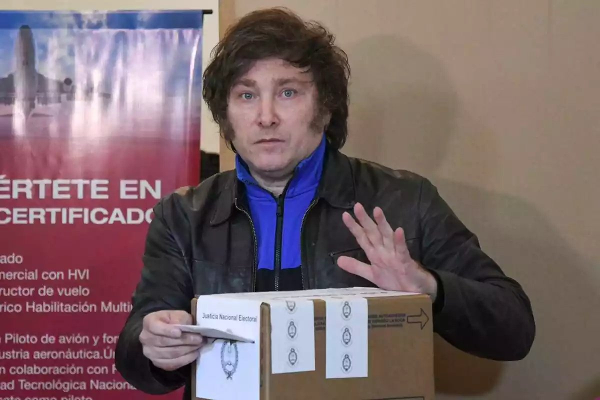 Pla mitjà de Javier Milei amb jaqueta de cuir introduint el seu vot en una urna de cartró i mirant fixament a càmera