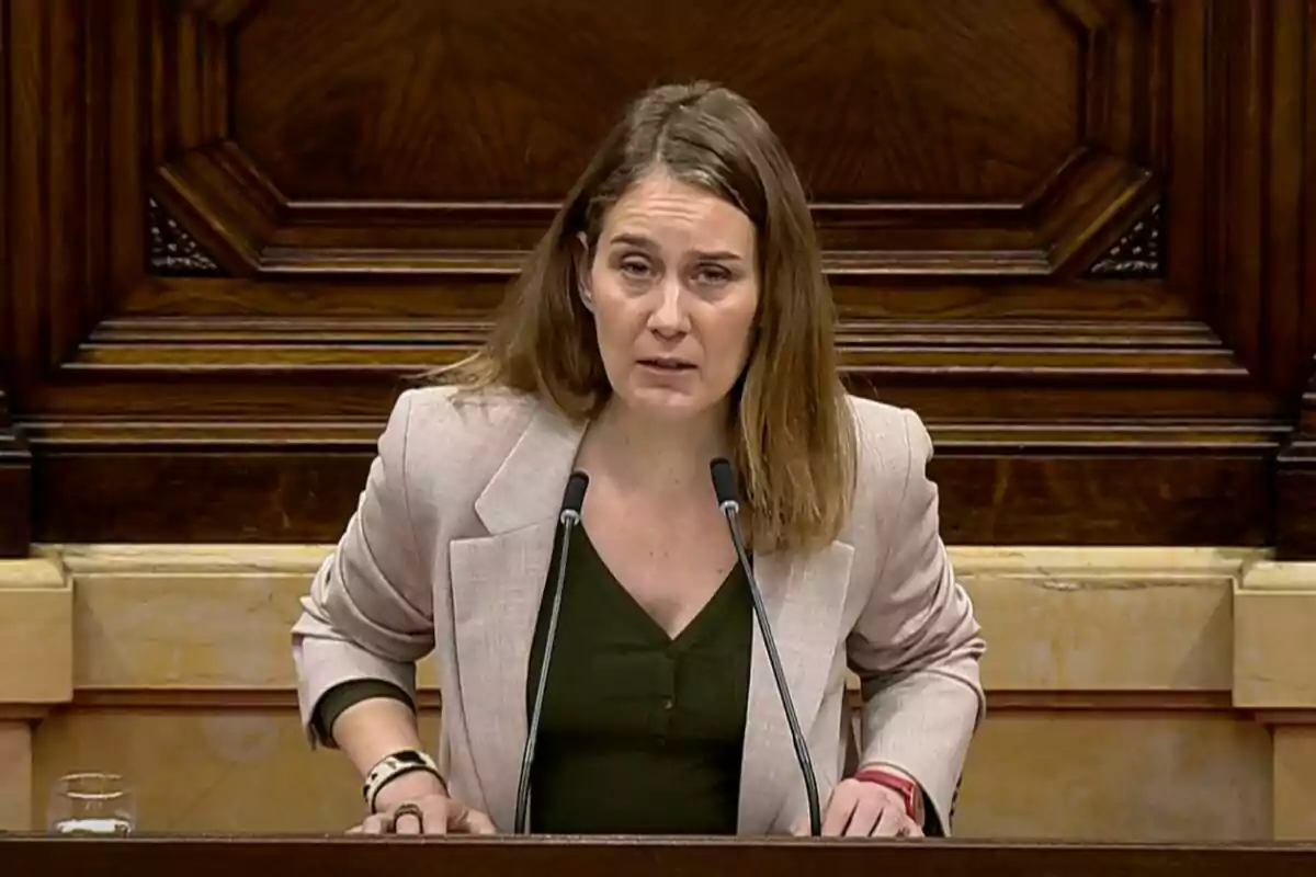 A woman speaking at a podium with two microphones in front of a wooden background. A woman speaking at a podium with two microphones in front of a wooden background.