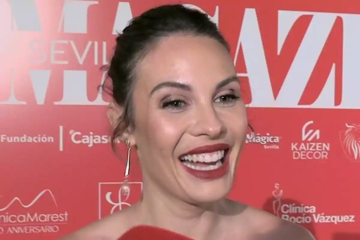 Jessica Bueno smiling in front of a red background with logos and text. Jessica Bueno smiling in front of a red background with logos and text.