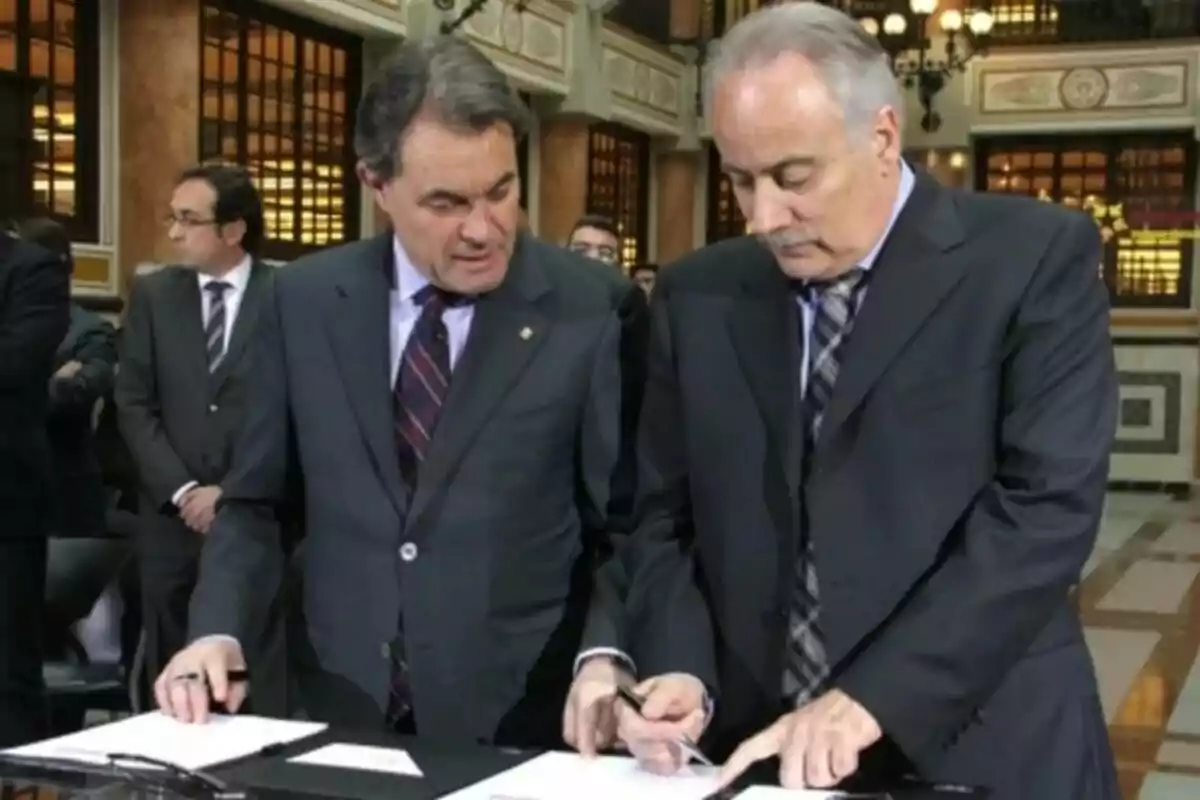 Dos homes amb vestit estan revisant documents en una taula en un entorn formal amb altres persones al fons.