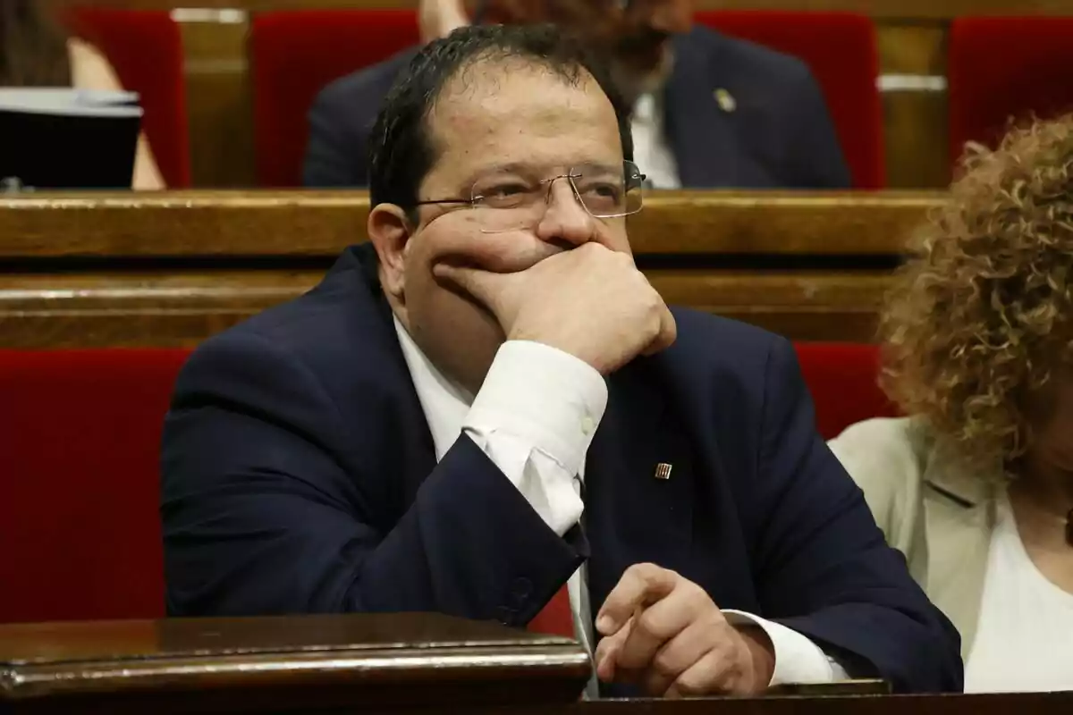 Joan Ignasi Elena està assegut al seu escó al Parlament, recolzant el seu cap a la mà i mirant pensatiu cap endavant.