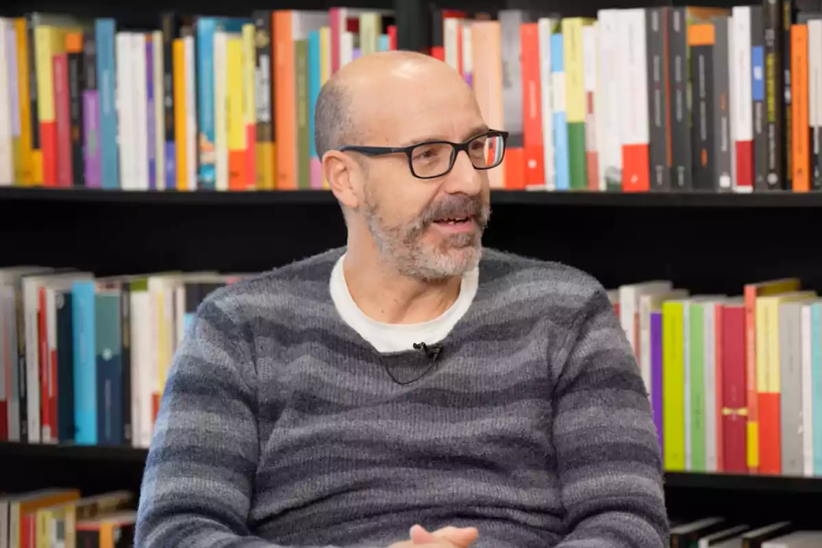 El periodista Joan Maria Pou sentado frente a una estantería llena de libros.