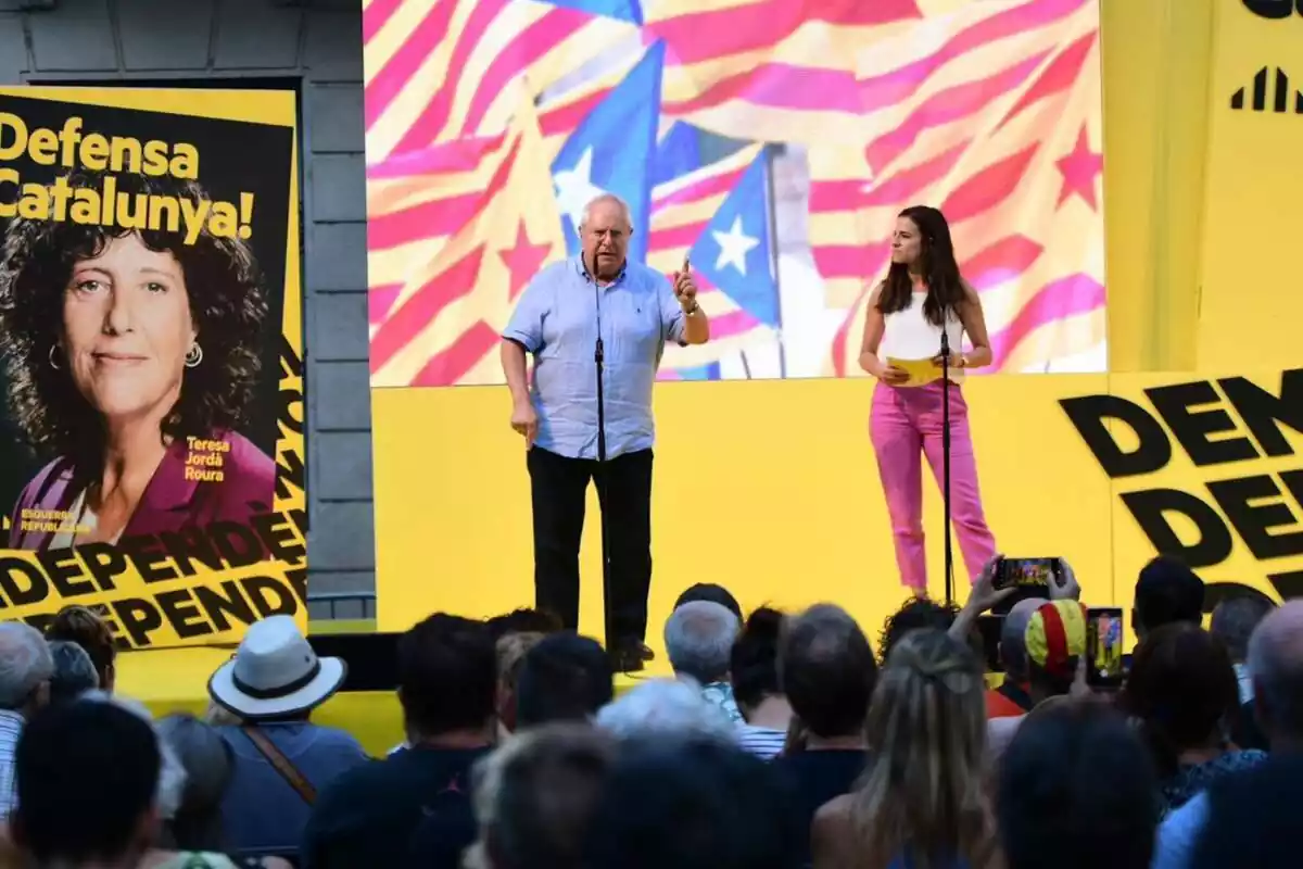 Pla general de Joan Queralt fent un míting davant de desenes de persones. De fons, una pantalla geganta groga amb imatges amb banderes estelades i un cartell electoral de Teresa Jordà