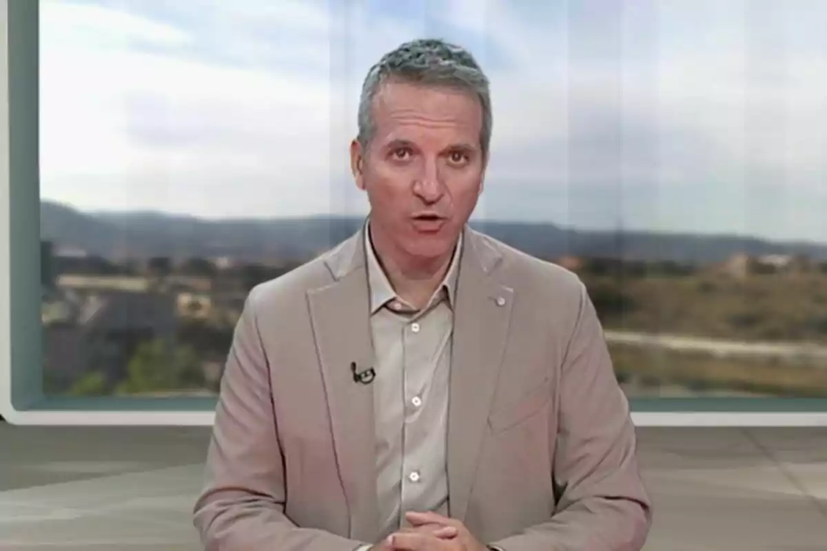 Pla mitjà de Jofre Janué vestit amb jaqueta beix parlant davant d’una càmera en un estudi amb un fons de paisatge difuminat.