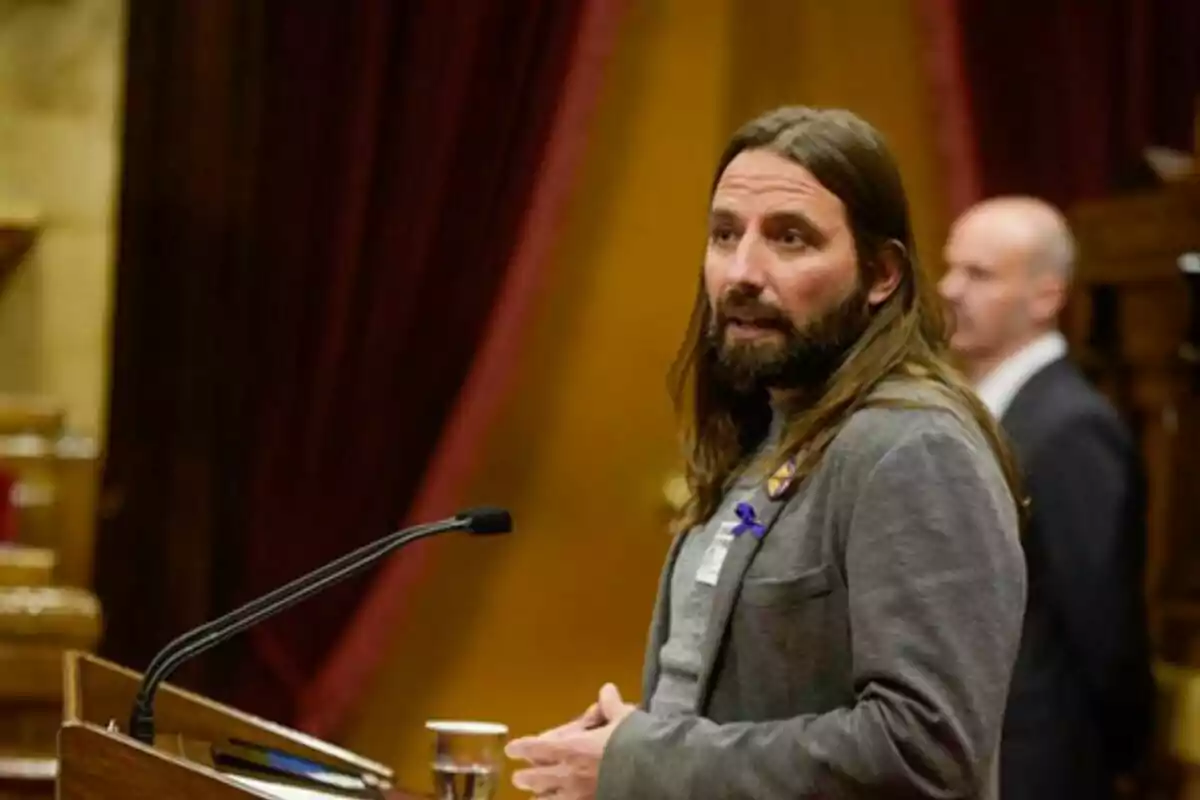 Un home amb cabell llarg i barba està parlant en un faristol en un entorn formal amb cortines vermelles al fons. Un home amb cabell llarg i barba està parlant en un faristol en un entorn formal amb cortines vermelles al fons.