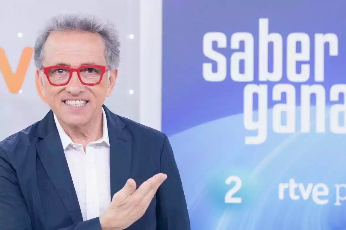 Jordi Hurtado de cabells canosos i ulleres vermelles somriu i assenyala amb la mà en un plató de televisió amb el logotip d’un programa anomenat Saber i Guanyar al fons.