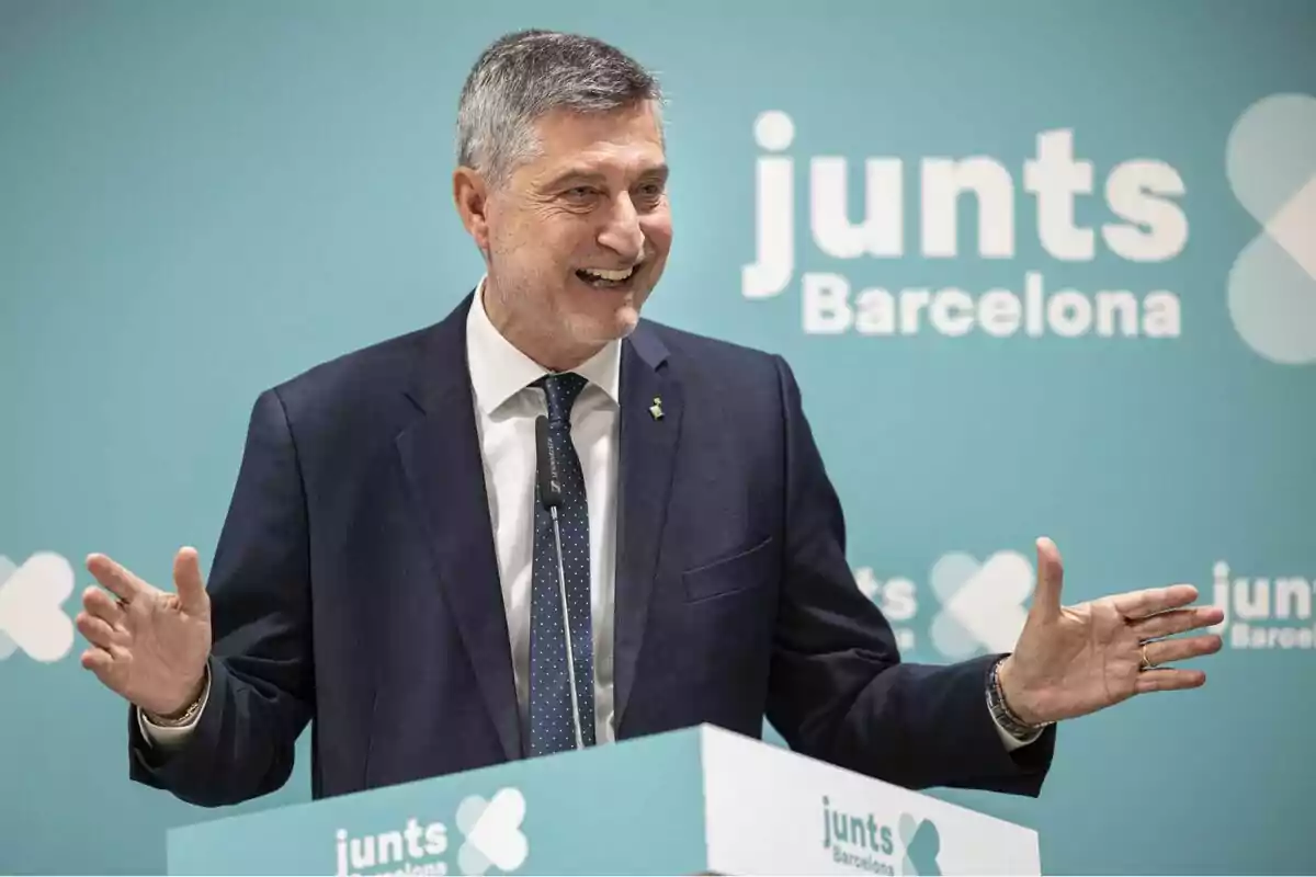 Un home amb vestit somriu mentre parla en un faristol amb el logotip de Un home amb vestit somriu mentre parla en un faristol amb el logotip de