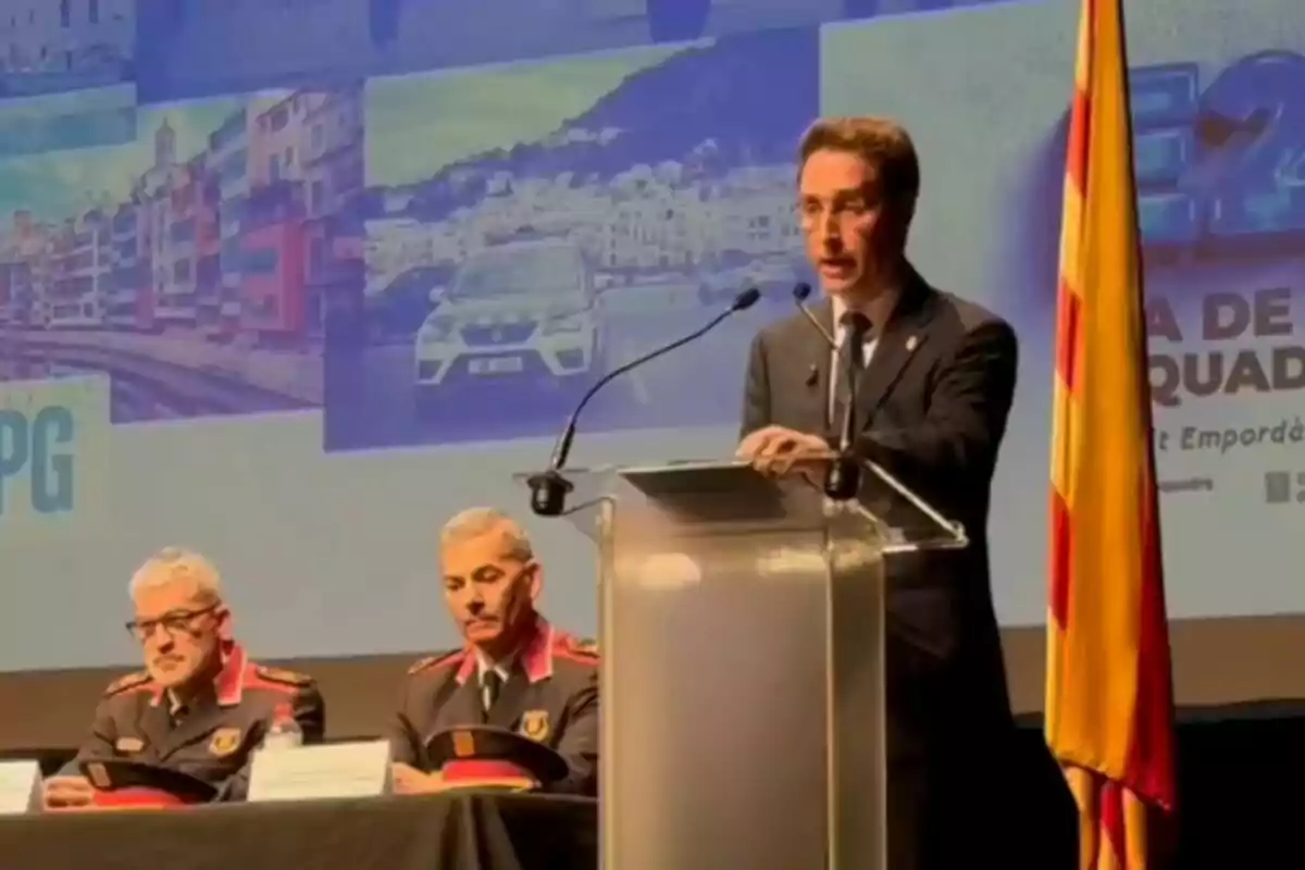 Un home en un podi parla en un esdeveniment oficial al costat de dos oficials uniformats asseguts, amb una bandera a la dreta i un fons amb imatges d'una ciutat i un cotxe.