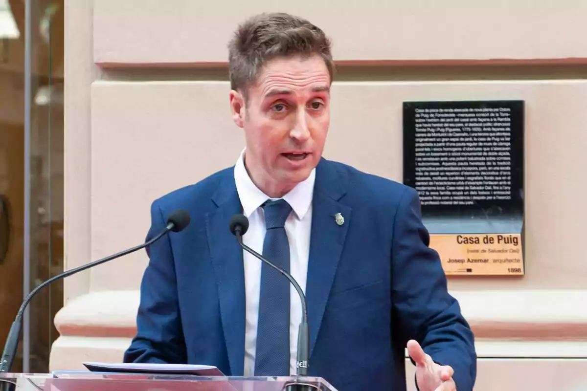 The mayor of Figueres, Jordi Masquef, in a blue suit speaking on a podium with two microphones in front of a commemorative plaque.