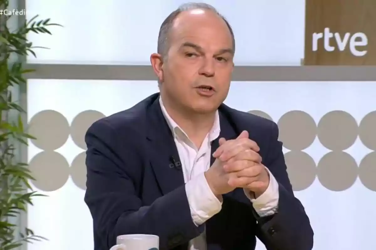 Un hombre con traje oscuro y camisa blanca está hablando en un set de televisión con el logo de RTVE visible al fondo.