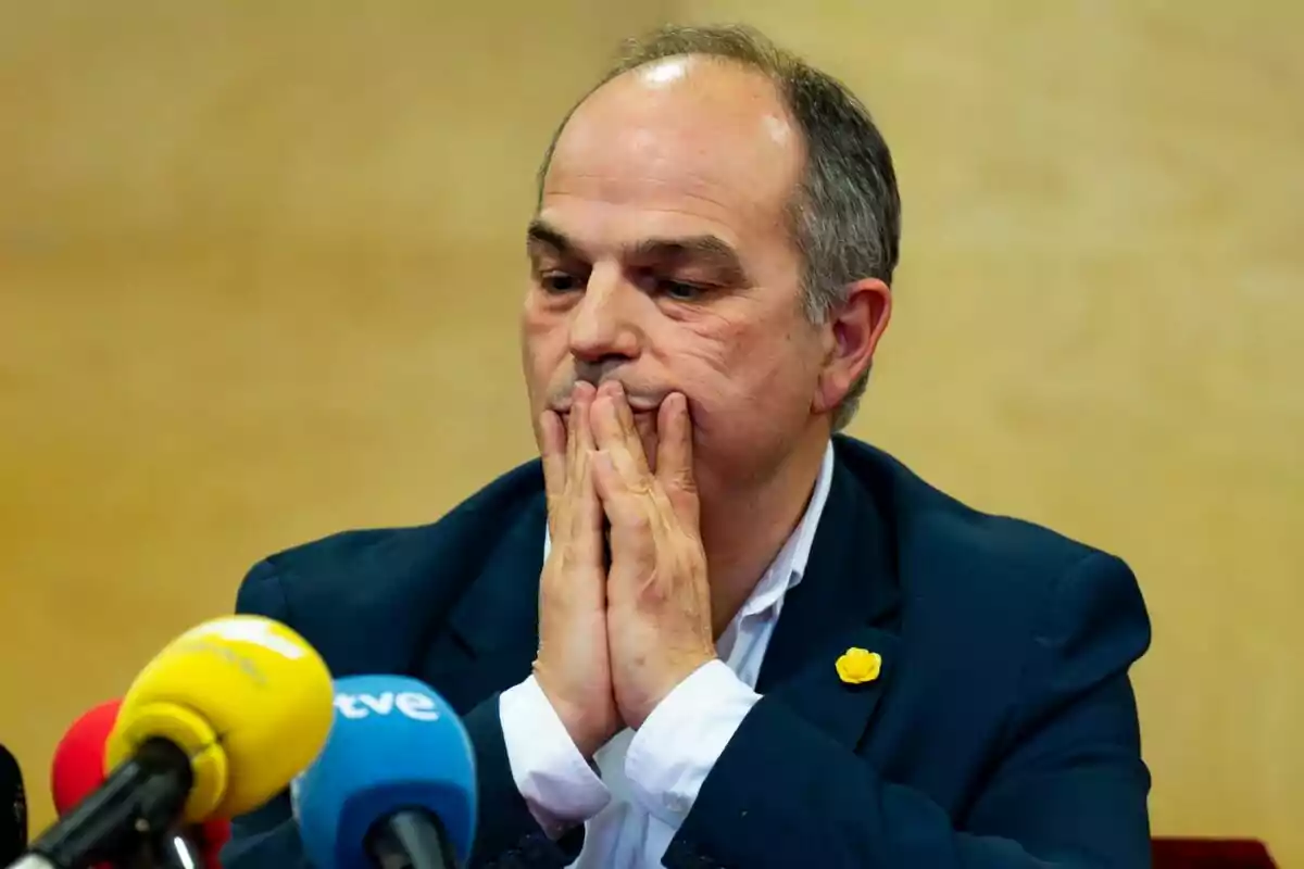 Un hombre con las manos juntas frente a su boca en una conferencia de prensa con micrófonos de colores en primer plano.