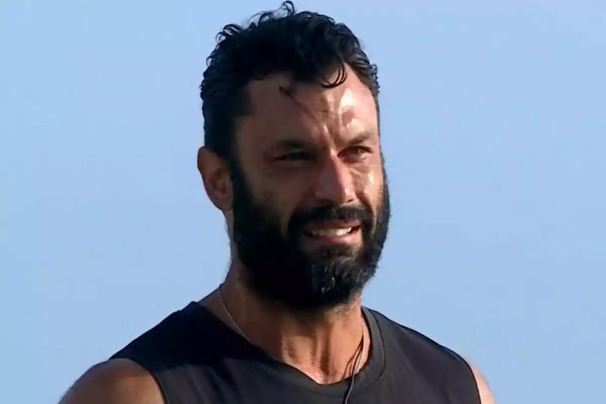 Fotografía de Jorge Pérez llorando con barba y cabello rizado, vistiendo una camiseta sin mangas negra, con el cielo azul de fondo en el puente de las emociones de Supervivientes