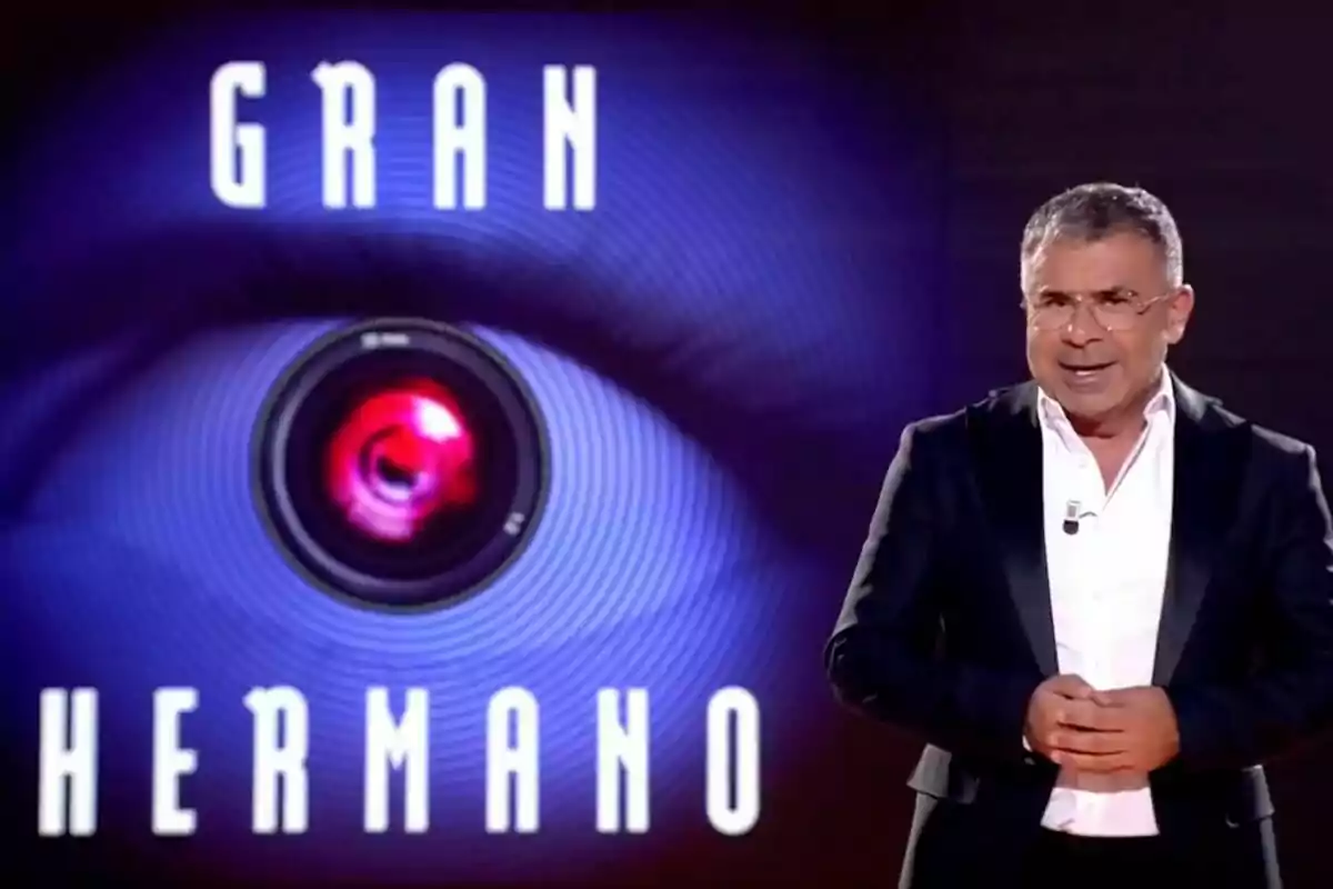 Jorge Javier Vázquez amb un vestit fosc i camisa blanca, al costat d'un logotip que diu "Gran Hermano" amb un ull i una càmera al centre.