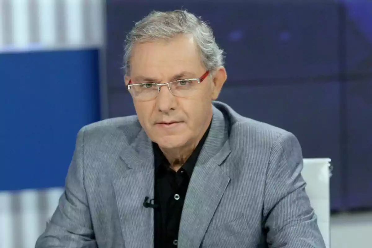 José Ángel de la Casa amb ulleres i cabell canós porta un vestit gris i camisa negra mentre està assegut en un estudi de televisió. José Ángel de la Casa amb ulleres i cabell canós porta un vestit gris i camisa negra mentre està assegut en un estudi de televisió.